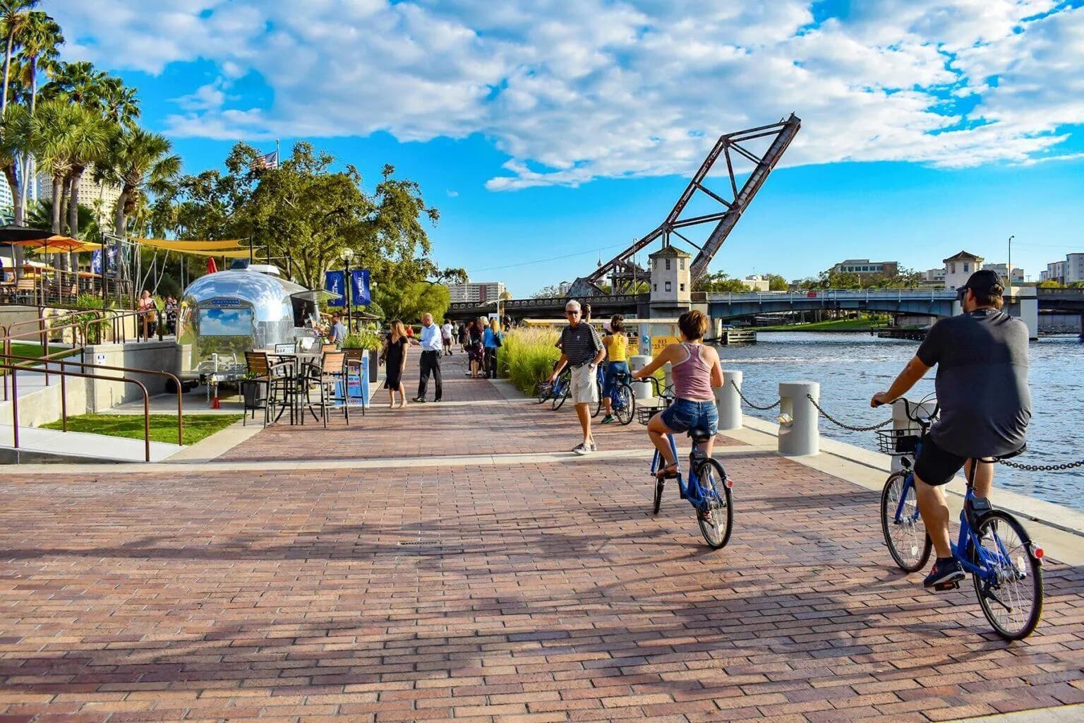 The Tampa Riverwalk today — cyclists and walkers on brick pavers, an Airstream cafe, the Cass Street bridge beyond