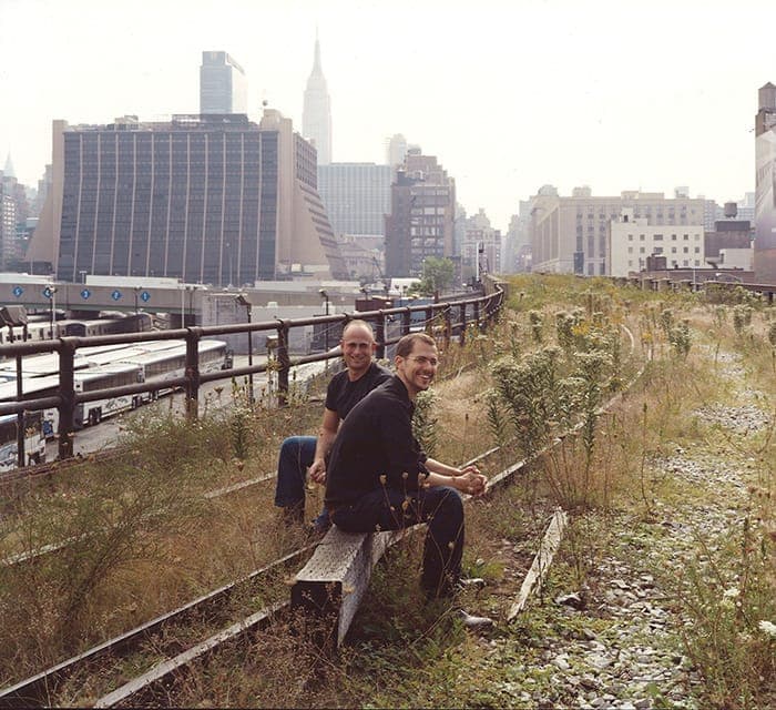 Robert Hammond and Joshua David sitting together on the High Line's abandoned tracks amid the wild growth, Midtown skyline behind, 1999