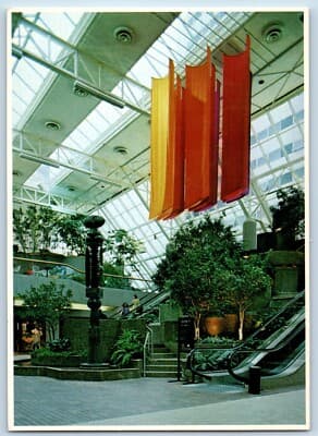 Postcard of Town Square Park, Saint Paul — orange and red banners hanging under the glass roof above trees, a dark sculptural fountain, and escalators